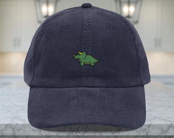 May include: Navy blue corduroy baseball cap featuring an embroidered green triceratops dinosaur. The cap has a curved brim and a structured crown. A casual accessory for everyday wear.