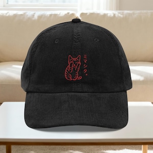May include: Black corduroy baseball cap with a red embroidered cat design and Japanese text. The cap has a curved peak and an adjustable strap. The cat illustration is in a minimalist style.