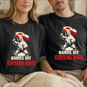 May include: Black t-shirts featuring a graphic of a white bear holding a Greenlandic flag. The bear is standing on a splash of white, with the words "HANDS OFF GREENLAND" in red and white text below the bear.