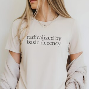 May include: A beige short-sleeved t-shirt featuring the black text "radicalized by basic decency" printed on the chest. The shirt has a crew neckline and is worn with a light-colored, textured, long-sleeved cardigan draped over the shoulders.