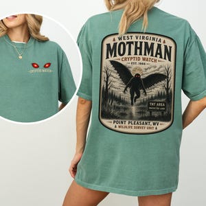 May include: A sage green t-shirt featuring a Mothman graphic on the back with the text West Virginia Mothman Cryptid Watch Est. 1966 Point Pleasant, WV Wildlife Survey Unit. The front chest area displays two glowing red eyes and Cryptid Watch text.