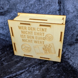 Könnte beinhalten: Eine hellfarbene Holzkiste mit einem Klappdeckel. Die Kiste hat eingravierten Text und Bilder von Euro-Währung. Der Text lautet "WER DEN CENT NICHT EHRT IST DEN EURO NICHT WERT".