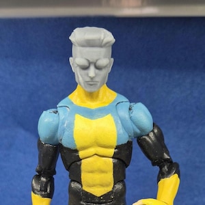 Puede incluir: Una figura de acción gris con un traje azul y amarillo. La figura tiene una cabeza esculpida con una expresión seria y un peinado corto y puntiagudo.