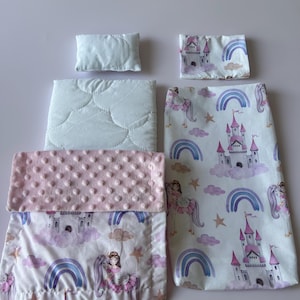 Op de afbeelding: Babyspeelgoedset met een klein kussen, een gewatteerde deken en een roze, getextureerde deken. De set heeft een prinsessen- en eenhoornthema met kastelen, regenbogen en sterren. Gemaakt van zachte stof.
