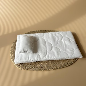 May include: A small, white quilted pet bed set with a matching pillow. The bed is rectangular with a wavy stitch pattern and sits on a round, woven mat. The background is a solid, light tan colour.