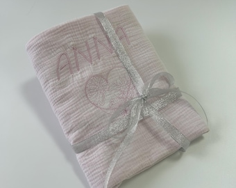 May include: A folded pink baby blanket with the name "ANNA" embroidered in pink thread. A heart-shaped design is also embroidered below the name. The blanket is tied with a silver ribbon.