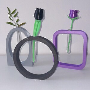 K&ouml;nnte beinhalten: Drei moderne geometrische Vasen in Grau, Schwarz und Lila, jede mit einem durchsichtigen Glasrohr zum Halten einer einzelnen Blume. Eine schwarze Tulpe, eine lila Rose und ein gr&uuml;ner Zweig sind in den Vasen ausgestellt.