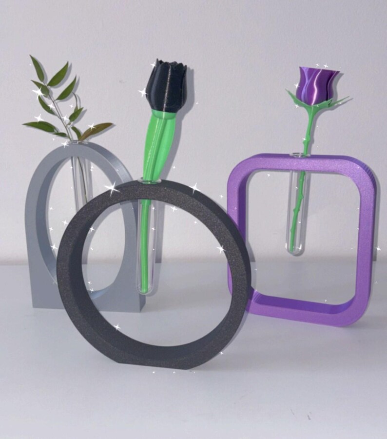 K&ouml;nnte beinhalten: Drei moderne geometrische Vasen in Grau, Schwarz und Lila, jede mit einem durchsichtigen Glasrohr zum Halten einer einzelnen Blume. Eine schwarze Tulpe, eine lila Rose und ein gr&uuml;ner Zweig sind in den Vasen ausgestellt.