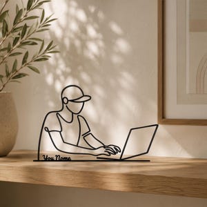 Peut inclure: Une sculpture en fil de métal noir représentant une personne portant une casquette et assise devant un ordinateur portable. La base de la sculpture comporte le texte "You Nome" en écriture cursive, intégré au design en ligne continue.