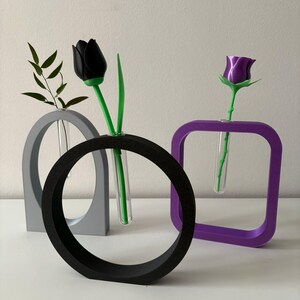 K&ouml;nnte beinhalten: Drei geometrische Vasen in Grau, Schwarz und Lila mit einer schwarzen Tulpe und einer lila Rose in den Vasen.