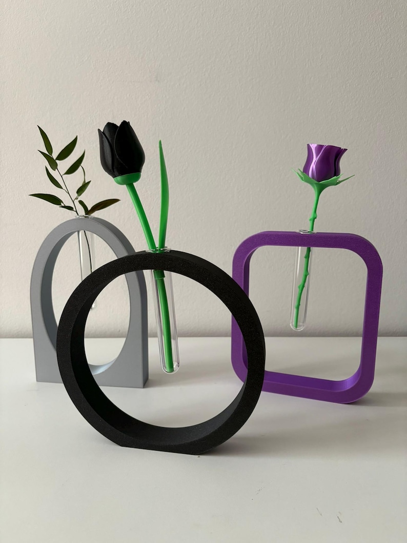 K&ouml;nnte beinhalten: Drei geometrische Vasen in Grau, Schwarz und Lila mit einer schwarzen Tulpe und einer lila Rose in den Vasen.