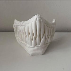Op de afbeelding: Een wit gezichtsmasker met een matte afwerking, voorzien van een sculpturaal ontwerp van scherpe, puntige tanden en hoektanden. Het masker bedekt het onderste deel van het gezicht en heeft een gestructureerd, stijf oppervlak met tanden.