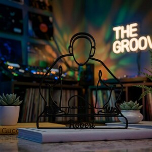 May include: Black metal DJ silhouette sculpture with headphones and a mixing console. The sculpture is placed on a book, with the name "Robert" at the base. The background features a neon sign that says "THE GROOVE".
