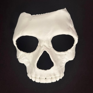 May include: A white, molded skull mask featuring large, hollow eye sockets and a triangular nasal cavity. The mask covers the upper portion of the face, extending from the forehead down to the upper teeth, with a smooth, matte finish throughout.