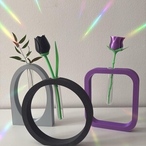 K&ouml;nnte beinhalten: Drei moderne geometrische Vasen in Grau, Schwarz und Lila mit klaren Glasr&ouml;hrchen zum Aufstellen von Blumen. Eine schwarze Tulpe und eine lila Rose werden in den Vasen ausgestellt.