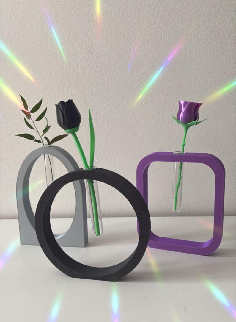 K&ouml;nnte beinhalten: Drei moderne geometrische Vasen in Grau, Schwarz und Lila mit klaren Glasr&ouml;hrchen zum Aufstellen von Blumen. Eine schwarze Tulpe und eine lila Rose werden in den Vasen ausgestellt.