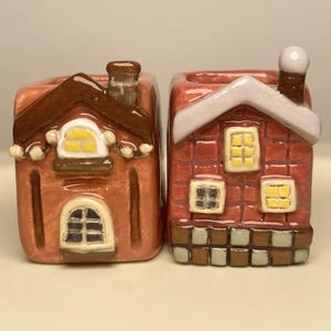 Puede incluir: Dos pequeñas casas de cerámica, una con techo marrón y bordes blancos, la otra con fachada de ladrillo rojo y techo blanco. Ambas casas tienen ventanas amarillas.