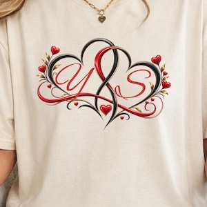 Puede incluir: Una camiseta de manga corta color crema con un diseño gráfico de dos corazones entrelazados en negro y rojo. El diseño incluye las letras Y y S en letra cursiva, rodeadas de pequeños corazones rojos y adornos decorativos.