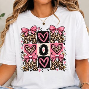 Könnte beinhalten: Ein weißes Kurzarm-T-Shirt mit einem zentralen Grafikdesign, das das Wort MOM in Leopardenmuster-Buchstaben zeigt, akzentuiert durch rosa Schleifen, rosa Herzen und kleine weiße Gänseblümchen, angeordnet in einem quadratischen Raster.