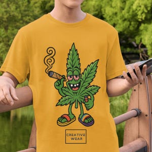 Può includere: T-shirt giallo senape con un personaggio di foglia di cannabis dei cartoni animati che fuma un sigaro e tiene un accendino. Il personaggio indossa sandali con cinturini arcobaleno. Il testo "CREATIVE WEAR" è in una casella in basso.