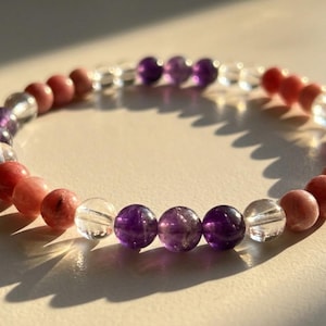Op de afbeelding: Een kralenarmband met amethist, heldere kwarts en roze rhodoniet edelstenen. De kralen zijn in een cirkelvormig patroon gerangschikt, waarbij de natuurlijke kleuren en texturen van de stenen worden getoond. De armband is ontworpen om om de pols te worden gedragen.