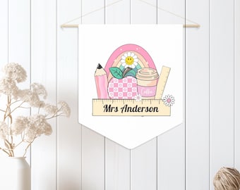 Pode incluir: Uma faixa de bandeira branca com um design temático de professor. O design inclui um lápis rosa, uma maçã rosa com um padrão xadrez, uma xícara de café, uma régua, um arco-íris e o texto "Mrs Anderson". A faixa é suspensa de um pino de madeira.