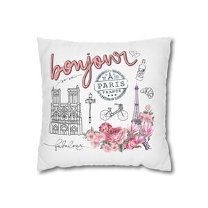 Könnte beinhalten: Weißes quadratisches Kissen mit rosa Schriftzug "bonjour" und Illustrationen zum Thema Paris. Mit Eiffelturm, Notre Dame, Fahrrad, Blumen und dem Wort "fabulous".