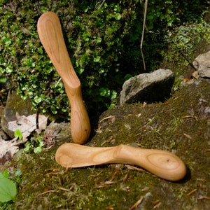Könnte beinhalten: Zwei Holzbuttermesser mit natürlicher Holzoptik. Die Messer liegen auf einem Bett aus grünem Moos und Steinen.