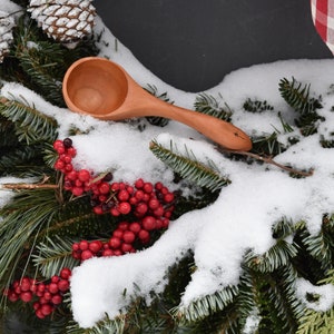 May include: A wooden scoop with a star design on the handle rests on a snowy evergreen wreath with red berries and pinecones. The scoop is made of light-colored wood and has a rounded bowl.