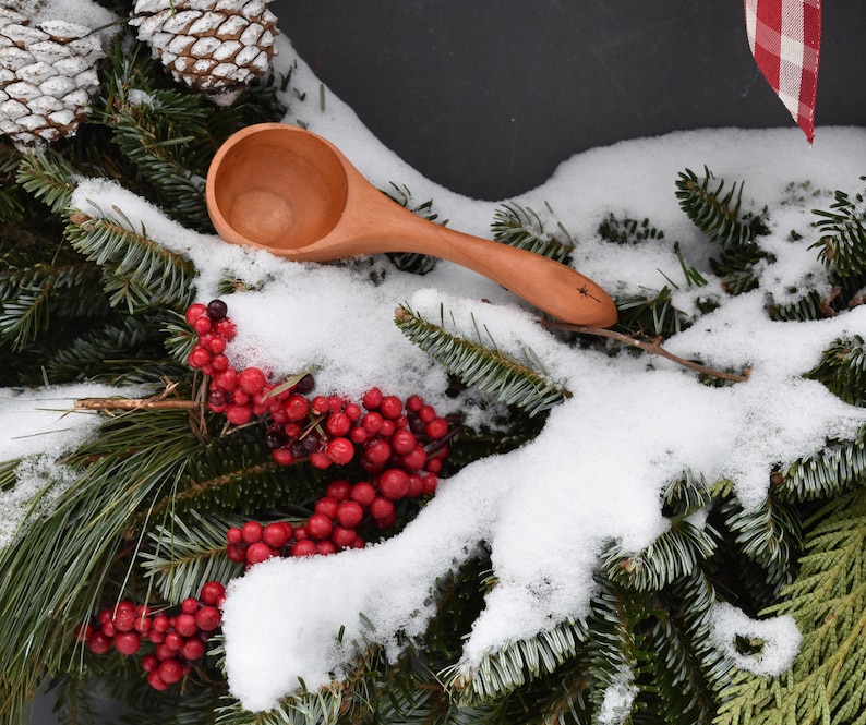May include: A wooden scoop with a star design on the handle rests on a snowy evergreen wreath with red berries and pinecones. The scoop is made of light-colored wood and has a rounded bowl.