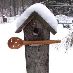 May include: A wooden birdhouse with a snow-covered roof, a wooden spoon with holes is attached to the front of the birdhouse.