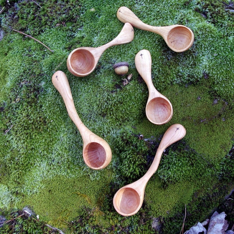 May include: Five wooden spoons with a light brown finish are arranged on a bed of green moss. Each spoon has a rounded bowl and a long, curved handle. A small acorn rests among the spoons. The spoons have a star design on the handle.