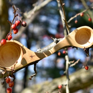 K&ouml;nnte beinhalten: Ein Holzl&ouml;ffel mit zwei Schalen, eine an jedem Ende, aus einem einzigen St&uuml;ck Holz geschnitzt. Der L&ouml;ffel liegt auf einem Ast mit roten Beeren.
