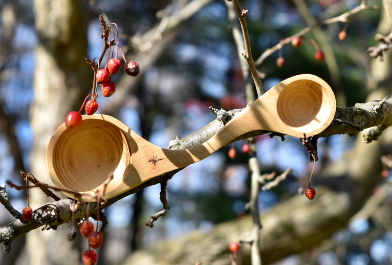 K&ouml;nnte beinhalten: Ein Holzl&ouml;ffel mit zwei Schalen, eine an jedem Ende, aus einem einzigen St&uuml;ck Holz geschnitzt. Der L&ouml;ffel liegt auf einem Ast mit roten Beeren.