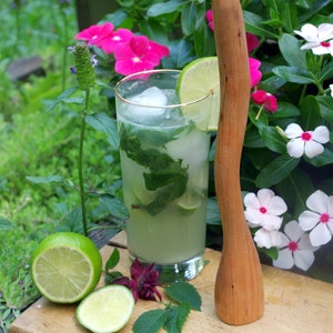 May include: A refreshing green mint mojito cocktail in a tall glass with ice, lime wedges, and fresh mint leaves. A wooden muddler sits on a wooden surface with pink and white flowers in the background.