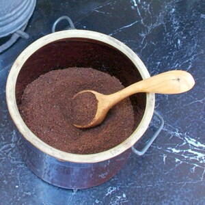 May include: A ceramic container filled with dark brown ground coffee, with a wooden scoop resting inside. The container has a light brown rim and two metal handles. The wooden scoop has a star design on the handle.
