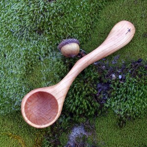 May include: A handcrafted wooden spoon with a rounded bowl and long handle, featuring a star-like carving at the end. The spoon is light brown and rests on a bed of green moss, next to a single acorn.