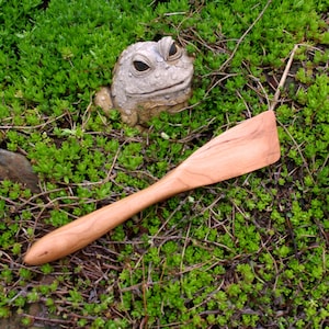May include: A wooden spatula with a long handle, made from cherry wood. The spatula is lying on a bed of green moss.