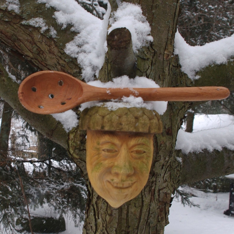 May include: A wooden carved face with a smiling expression is attached to a tree trunk. The face is covered in snow and a wooden spoon with holes is placed on top of the face.