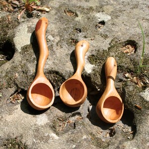 May include: Three handcrafted wooden spoons with curved handles and rounded bowls. The spoons are made of light-colored wood, with a natural finish. Each spoon has a unique shape and a small decorative element on the handle.