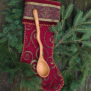 May include: A wooden spoon with a curved handle sits inside a red stocking with gold trim. The stocking is surrounded by green pine branches.