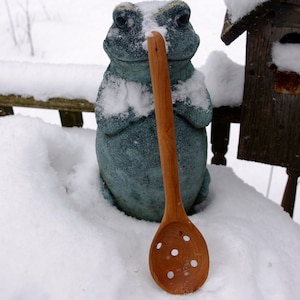 May include: A green frog statue with a wooden slotted spoon sticking out of its mouth. The frog is sitting in a snowy scene.