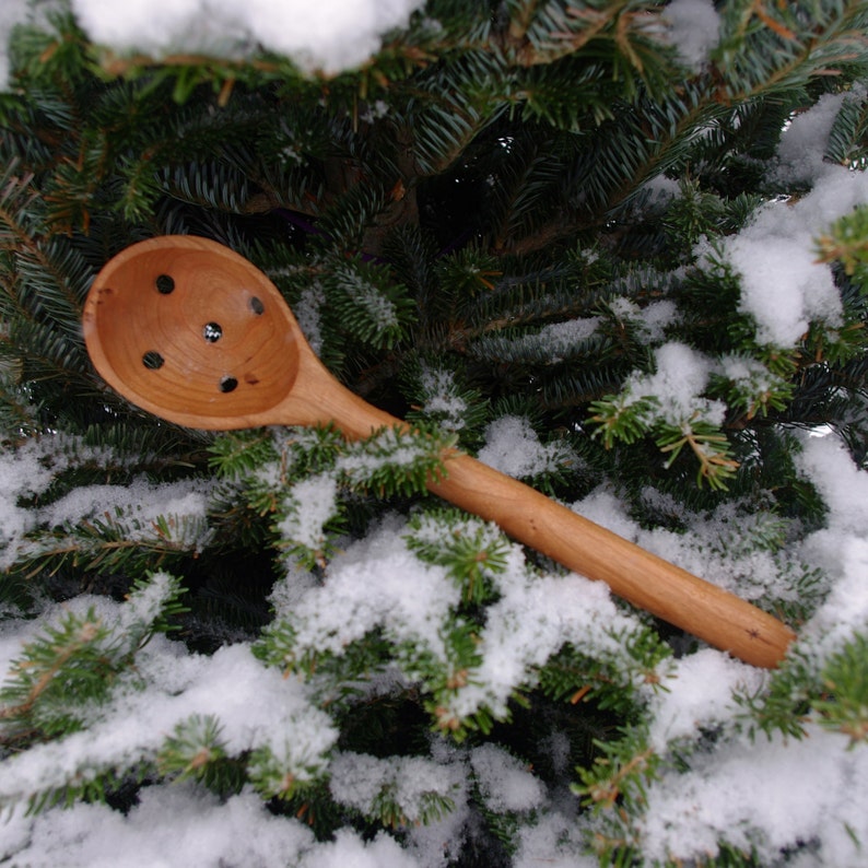 May include: A wooden spoon with holes in the bowl, resting on a snowy evergreen tree branch.