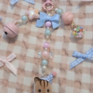 May include: A keychain featuring a flocked animal figure wearing a blue and white checkered outfit and hat. The charm is attached to a pink heart-shaped clasp and decorated with pastel beads, a blue bow, a pink bell, and a clear sphere with colorful balls.