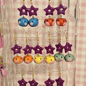 May include: A display of handmade earrings with gold-colored hooks. Each pair features a purple star charm and a unique, colorful bead. Bead designs include blue with white dots, orange with white accents, and pink with black details.