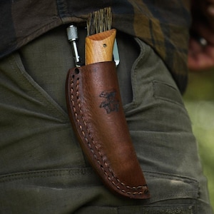 Può includere: Un fodero in pelle marrone contenente un coltello per funghi con manico in legno e una spazzola all'estremità. Il fodero presenta un disegno di fungo stampato e cuciture visibili lungo i bordi. Un piccolo utensile in metallo è inserito lateralmente.