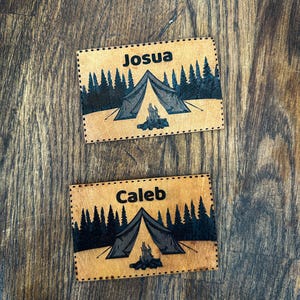 Può includere: Due toppe rettangolari in pelle con disegni a tema campeggio. Ogni toppa presenta una tenda, un falò e una silhouette di foresta. Una toppa recita "Josua" e l'altra "Caleb" in nero. Le toppe sono su una superficie di legno.