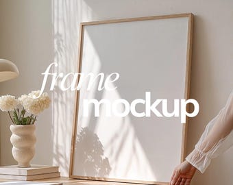 Può includere: Un mockup di stampa artistica con cornice in legno con il testo "frame mockup" in bianco. La cornice è appoggiata a un muro, con una mano che tiene la parte inferiore della cornice. Un vaso di fiori e libri sono su un tavolo di legno.