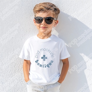 May include: A white t-shirt with a circular logo that reads "pod mockups boutique" in a light blue color. The t-shirt is being worn by a person wearing black sunglasses and light-colored jeans.