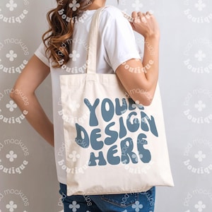 May include: Cream-colored tote bag with the text "YOUR DESIGN HERE" in blue, held by a person. The bag has a simple design, with a rectangular shape and long handles. The text is in a retro font.